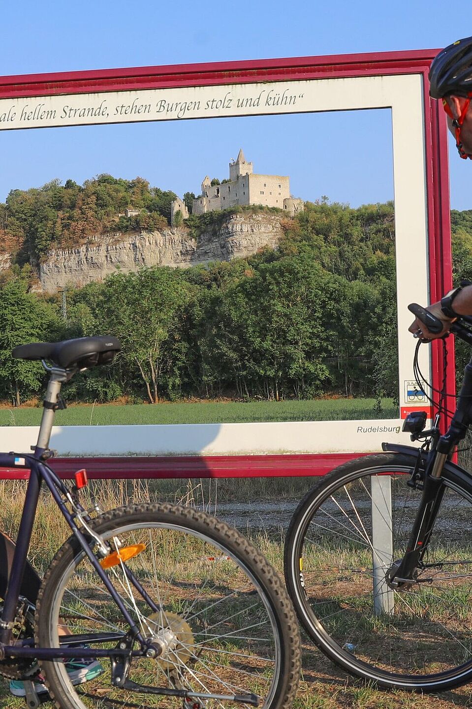 Die Rudelsburg an der Saale Eine Radfahrerin und ein Radfahrer stehen vor einem Bilderrahmen, durch den die Rudelsburg an der Saale zu sehen ist.
