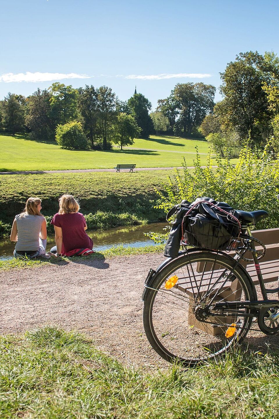 Der Ilmtal-Radweg führt durch den Schlosspark in Tiefurt ZWei Radfahrende sitzen an der Ilm im Schlosspark Tiefurt.