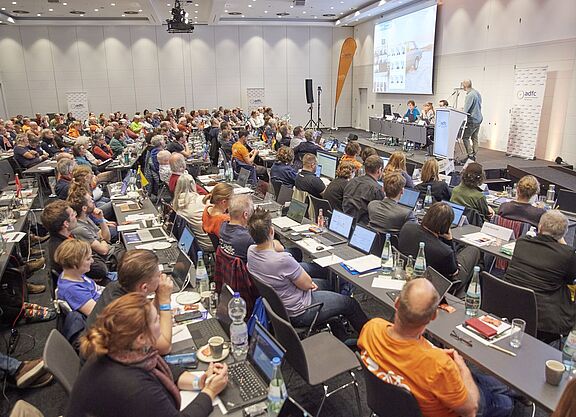 Ein grpßer Saal mit 140 Menschen an Tischreihen, die zur Bühne blicken, wo Boris von Heesen einen Vortrag hält. 